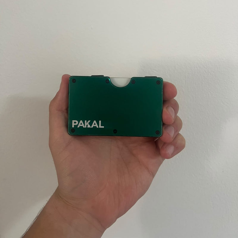 A hand holding a small green metal card case with the brand name 'PAKAL' printed in white on the front, set against a plain off-white wall.